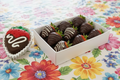 A white gift box filled with chocolate-covered strawberries, with one strawberry placed outside the box on the left. The strawberries are dipped in dark chocolate and decorated with drizzles of white chocolate, with green leafy tops visible.